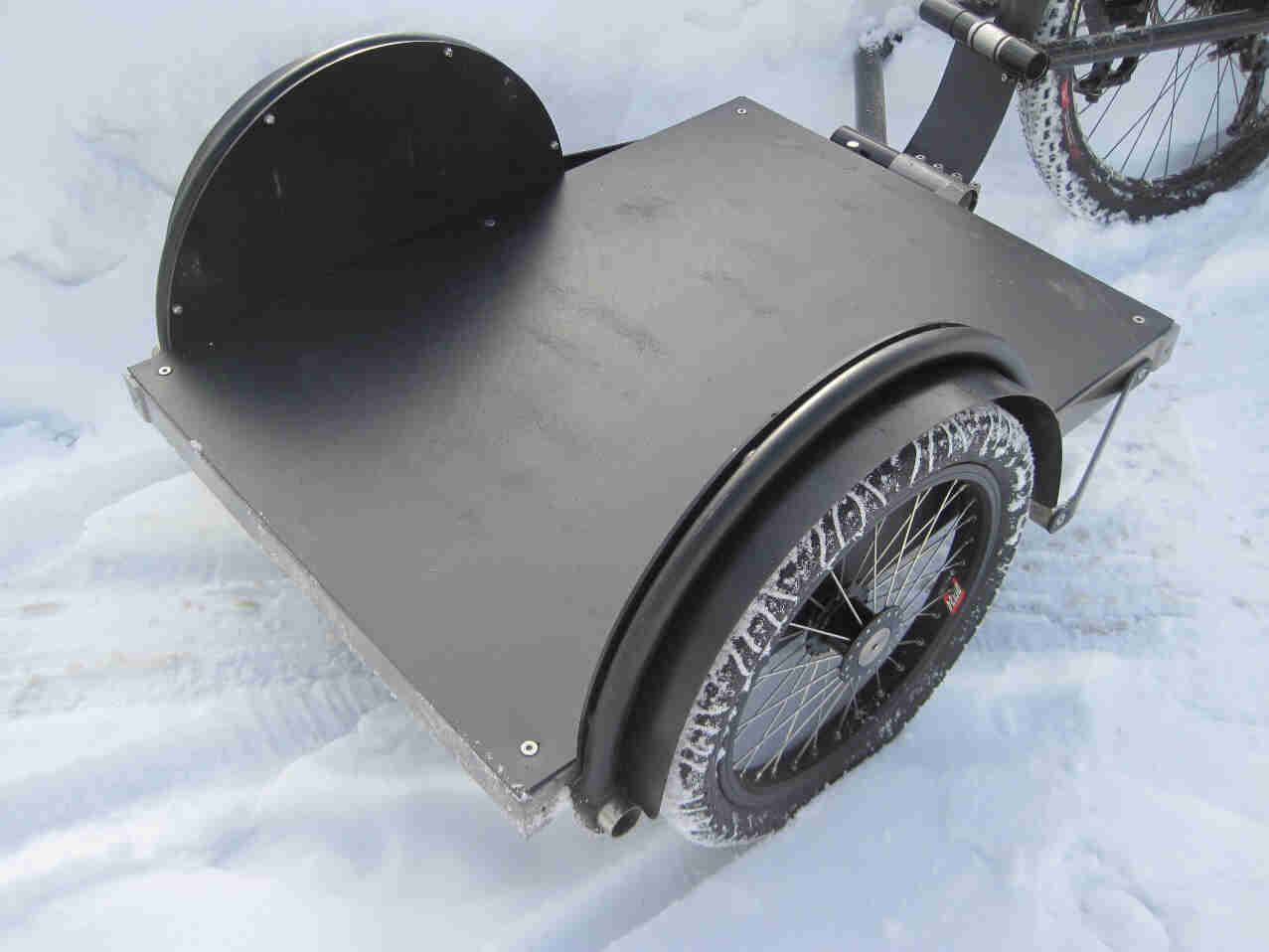 Downward, rear, right side view of a Surly Ted bike trailer with a flat top, parked on snow covered ground